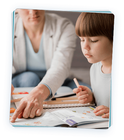 A parent guides a young child through a workbook activity, pointing to illustrations as the child writes with a pencil during a learning session.