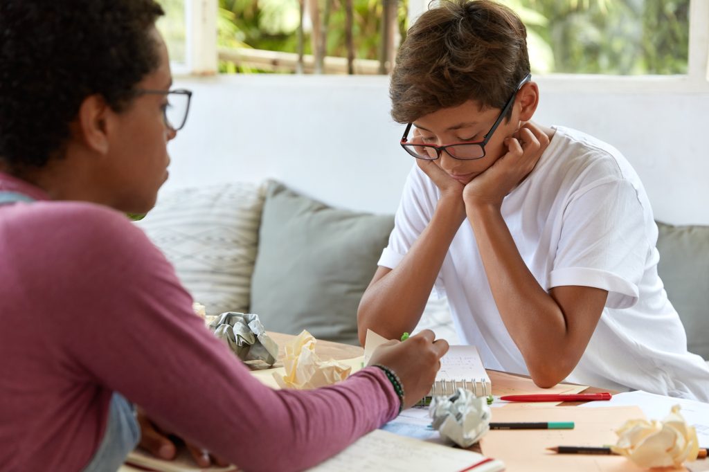 Adolescent portant des lunettes, visiblement découragé devant ses notes froissées, accompagné d'un adulte lors d'une évaluation neuropsychologique.