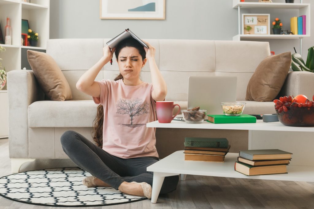 Jeune femme frustrée tenant un livre sur sa tête, entourée de livres et d'un ordinateur, illustrant les difficultés de la déficience intellectuelle légère.