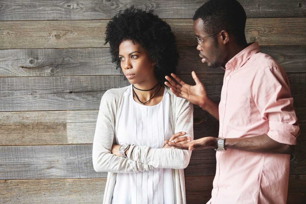  Femme aux bras croisés, détournant le regard, appuyée contre un mur de bois; un homme en chemise rose lui parle, la main tendue.