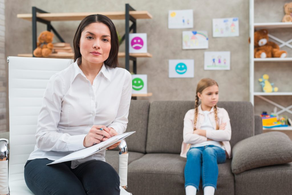  Intervenante avec une planchette, assise devant une fillette aux bras croisés, lors d’une évaluation neuropsychologique de l’enfant.