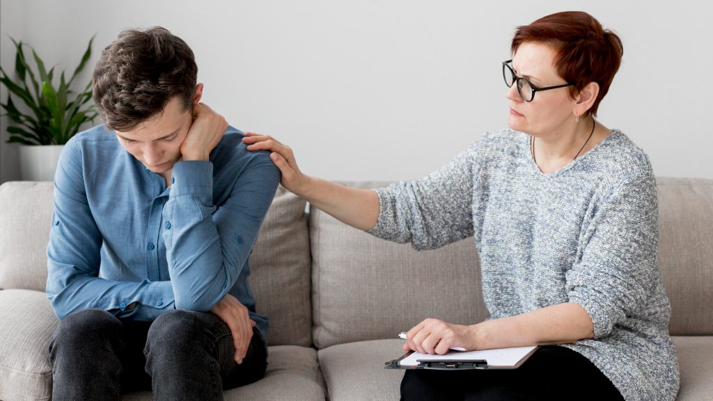 Professionnelle tenant un clipboard et posant une main bienveillante sur l'épaule d'un jeune adulte lors d'une évaluation neuropsychologique du TSA.