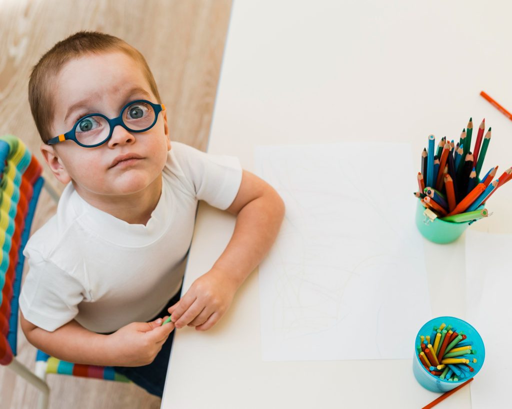 Jeune enfant avec trisomie 21 portant des lunettes rondes, regardant vers le haut près d'une table avec des crayons de couleur.