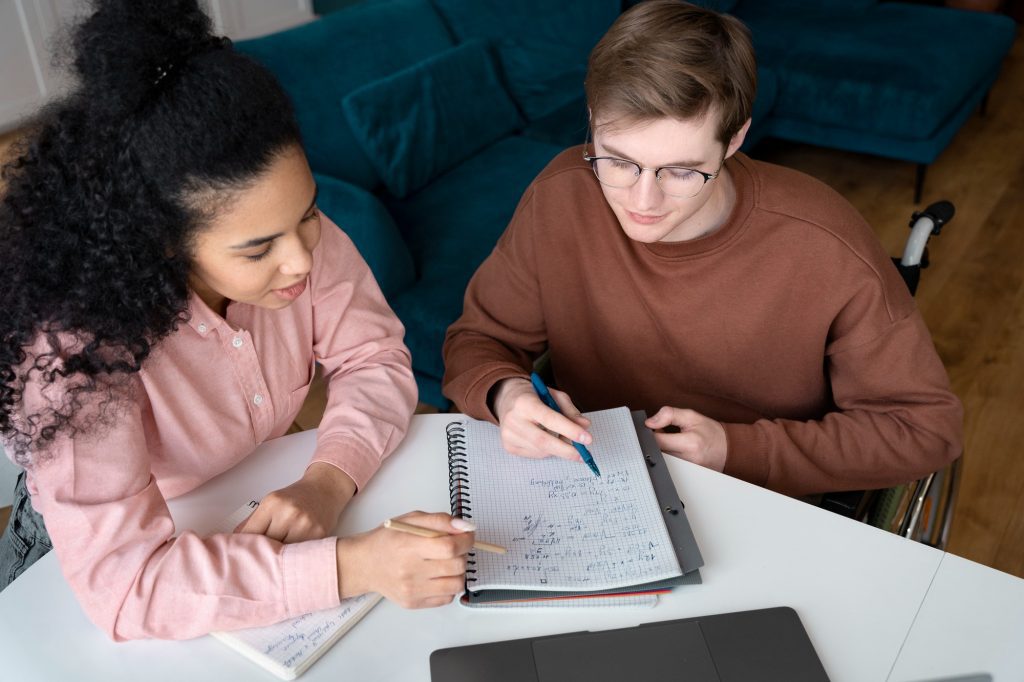 Deux jeunes adultes dont une personne en fauteuil roulant travaillant ensemble sur un cahier, illustrant le soutien après un diagnostic autisme adulte.