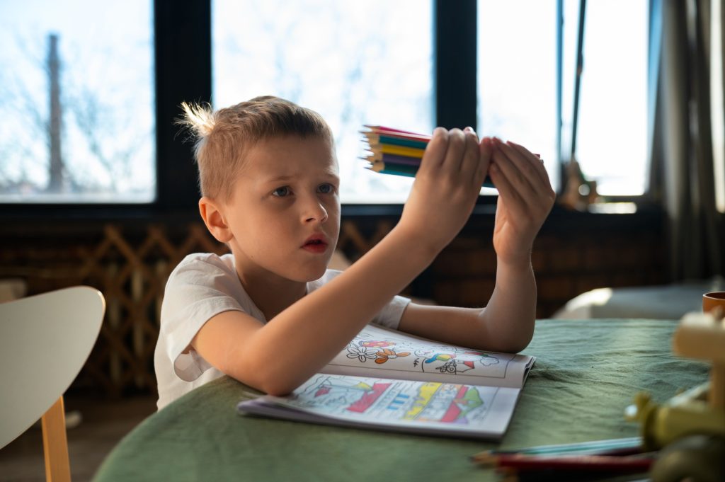 Garçon concentré tenant des crayons de couleur en éventail, avec un cahier ouvert devant lui, illustrant les difficultés cognitives liées à la trisomie 21.