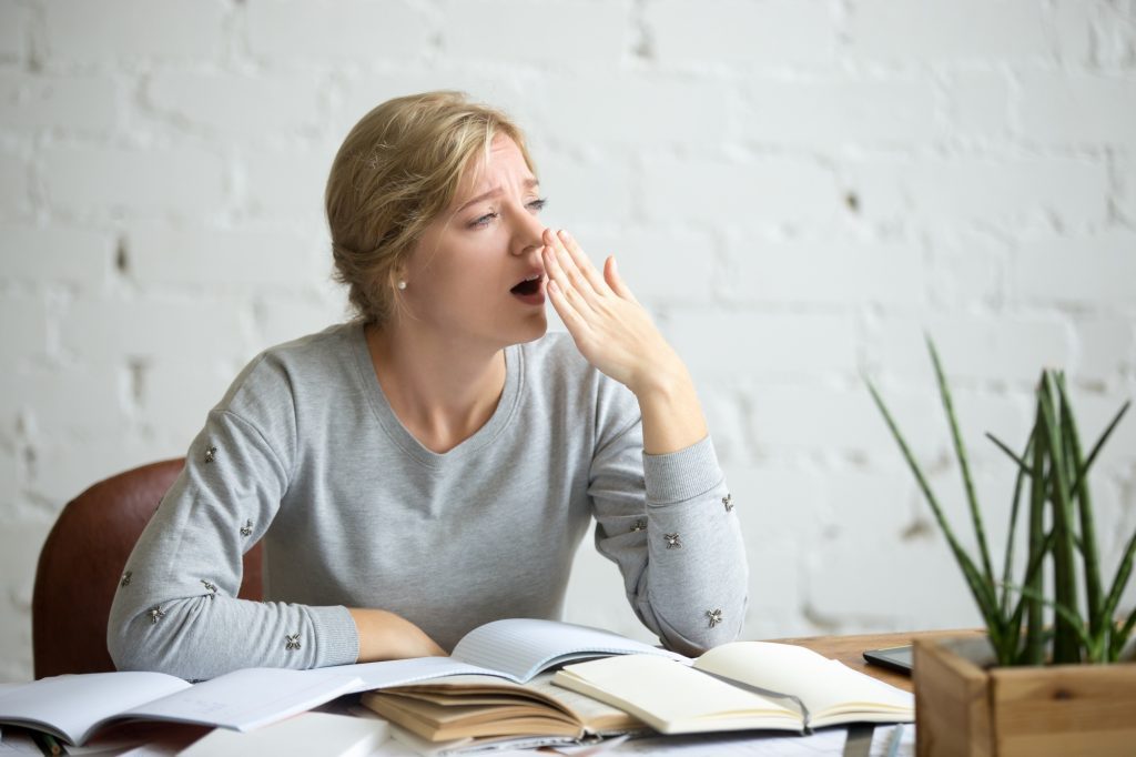 Jeune femme fatiguée à son bureau, le bureau couvert de livres ouverts, la main devant la bouche, épuisée par la charge cognitive au quotidien. 