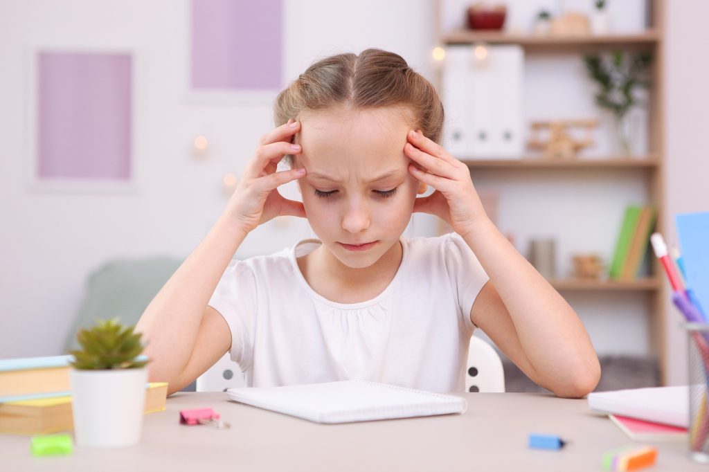 La fillette se tient la tête, dépassée à son bureau, reflet de difficultés liées au TDAH ou au TSA.