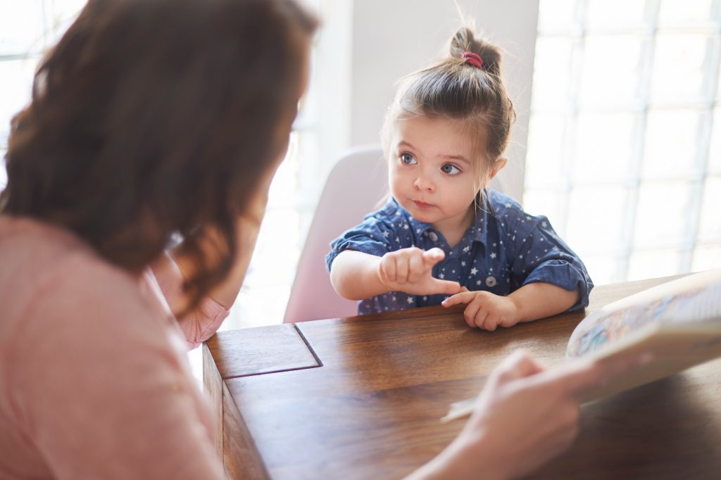 Petite fille pointant du doigt un livre illustré, assise à table avec un adulte lui faisant la lecture.